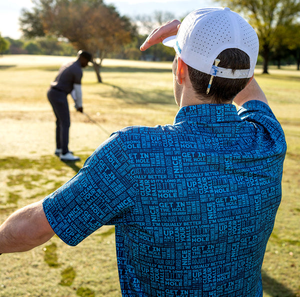 A man wearing the That's What She Said - Polo in an athletic fit and a white cap stands with his back to the camera, shading his eyes as he watches another golfer prepare to hit a ball on a sunny, green course.