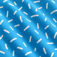 A close-up of folded Bitty Bombs - Women’s Polo fabric in vibrant blue, featuring a repeating pattern of small, white, slightly curved dash marks or sprinkles scattered across the surface. The material has a visible woven texture and offers UPF 50 protection, while the folds create subtle shadows that add depth to the polo’s bright blue appearance.