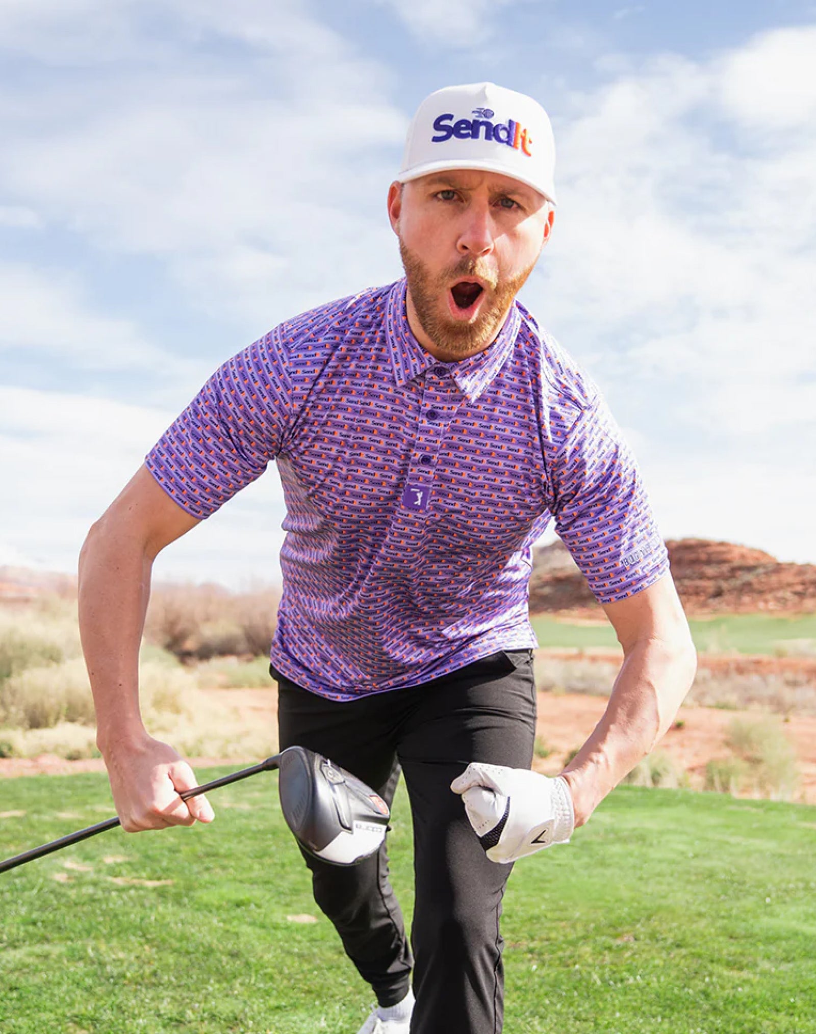 A bearded man wearing a Send it hat, a purple patterned polo, black pants, and white gloves, stands on a golf course holding two golf clubs, leaning forward with an excited facial expression, mouth open, on a sunny day with blue sky and rocky landscape.