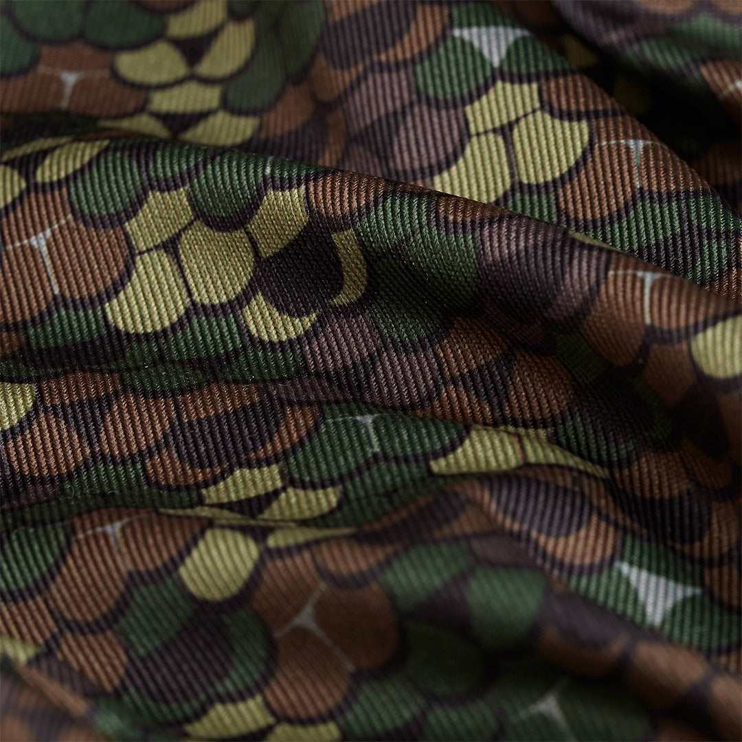 A close-up of the Whale Tail - Camo - Polo shows scalloped, scale-like patterns in green, brown, and tan. The ribbed, moisture-wicking fabric adds depth and comfort, with a camouflage look reminiscent of reptile skin.